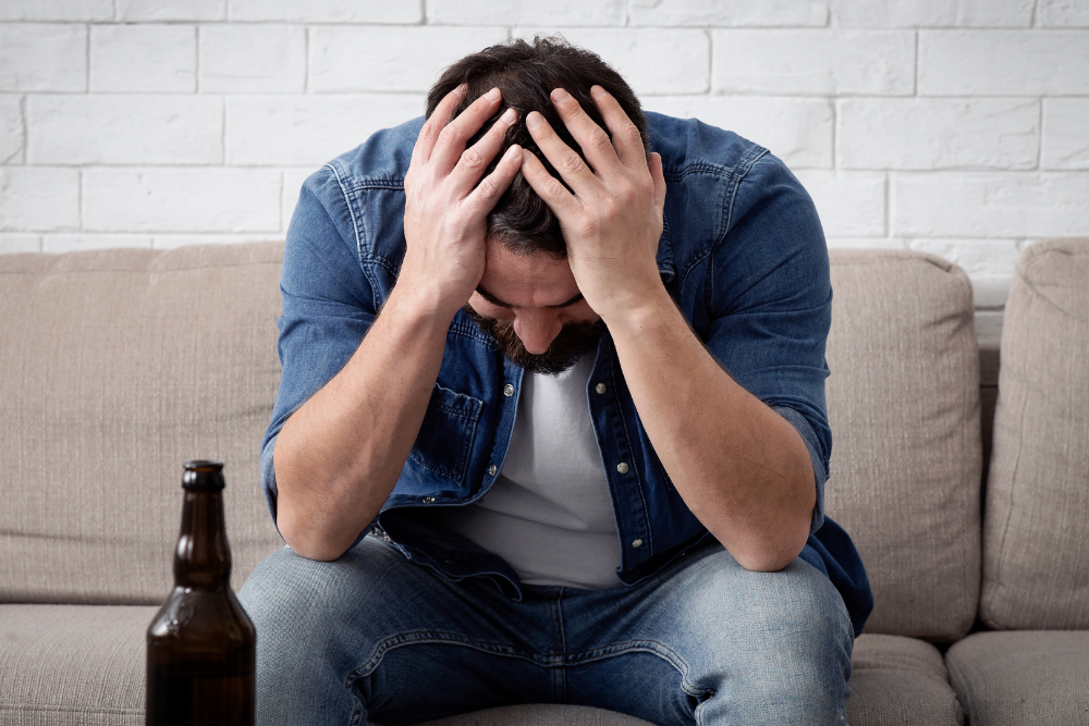 Distressed man sitting on a couch with a beer bottle, showing signs of depression and substance abuse.