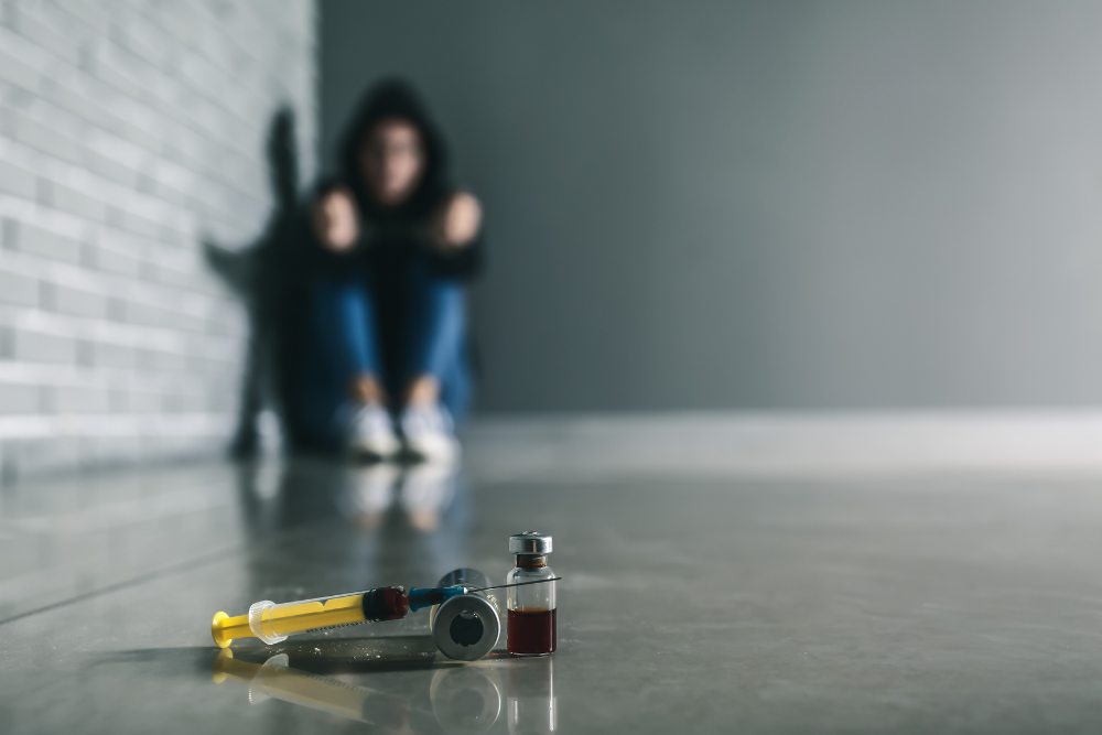 Syringes and drug vial on the floor with blurred figure behind, symbolizing substance abuse and mental health struggles.